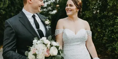 A bride in a white lace gown and a groom in a dark suit holding a bouquet of white and pink flowers, smiling at each other outdoors.