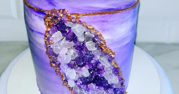 A tall, purple-frosted custom cake decorated with a realistic crystal geode design and gold accents.