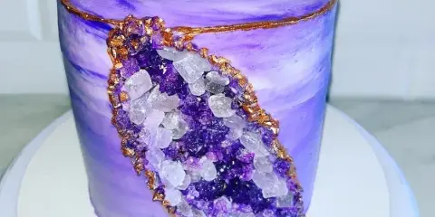 A tall, purple-frosted custom cake decorated with a realistic crystal geode design and gold accents.
