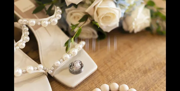 White pearl-embellished bridal heels, white rose bouquet, and an engagement ring on a wooden surface.