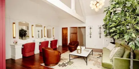 Interior view of a venue lounge featuring a green sofa, leather armchairs, a vanity with mirrors, and a large indoor tree.