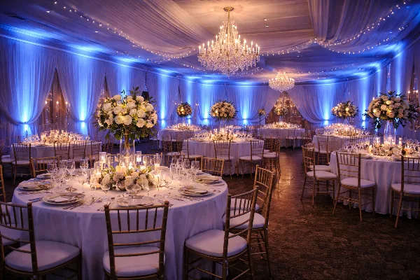 A wedding banquet hall with round tables, chandeliers, and blue uplighting illuminating the white drapery.