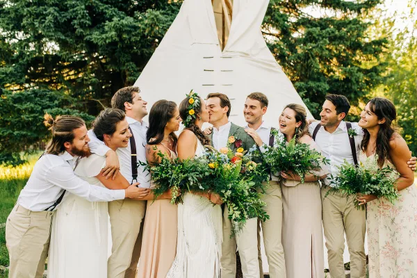 A wedding party group photo featuring a bride and groom kissing, surrounded by smiling bridesmaids and groomsmen holding bouquets, with a white teepee structure in the background.