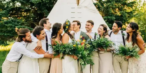 A wedding party group photo featuring a bride and groom kissing, surrounded by smiling bridesmaids and groomsmen holding bouquets, with a white teepee structure in the background.