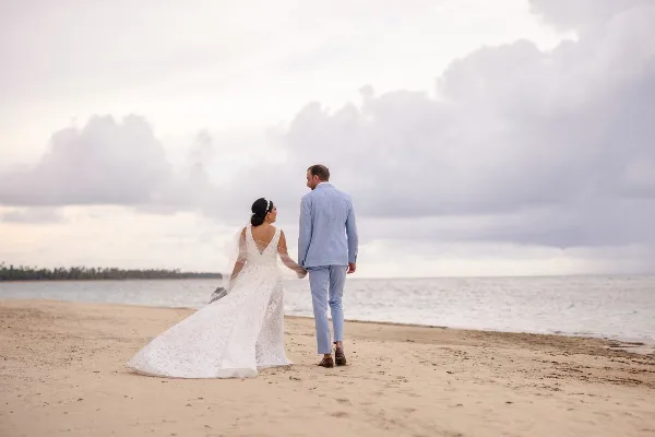 A bride and groom walking hand-in-hand on a sandy beach with the ocean and cloudy sky in the background.