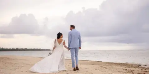 A bride and groom walking hand-in-hand on a sandy beach with the ocean and cloudy sky in the background.