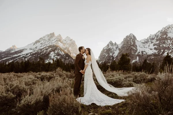 A bride and groom standing in a field with snow-capped mountains in the background.