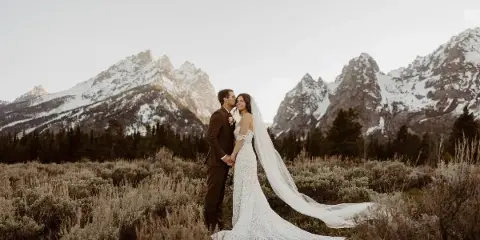 A bride and groom standing in a field with snow-capped mountains in the background.