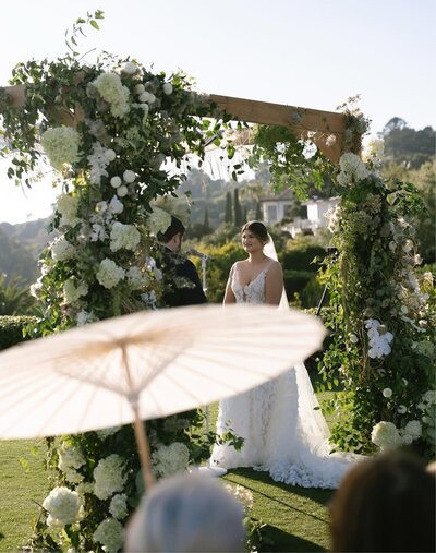 A bride in a white wedding dress stands beneath a large wooden archway decorated with lush greenery and white flowers during an outdoor ceremony.