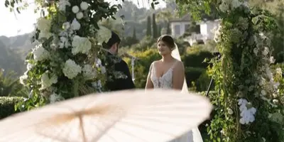 A bride in a white wedding dress stands beneath a large wooden archway decorated with lush greenery and white flowers during an outdoor ceremony.