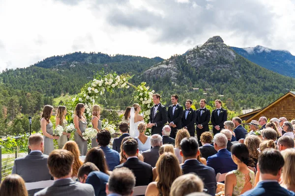 A wedding ceremony taking place outdoors with a bride and groom at an altar, surrounded by a wedding party and seated guests, with a mountain landscape in the background.