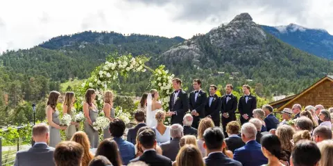 A wedding ceremony taking place outdoors with a bride and groom at an altar, surrounded by a wedding party and seated guests, with a mountain landscape in the background.