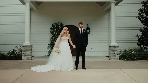 Bride in a white gown and groom in a black suit holding hands on a porch with floral arrangements.