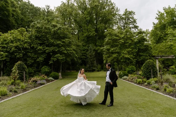A bride in a white gown and a groom in a tuxedo standing on a grassy path in a lush garden.
