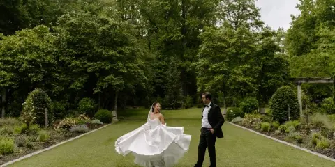 A bride in a white gown and a groom in a tuxedo standing on a grassy path in a lush garden.