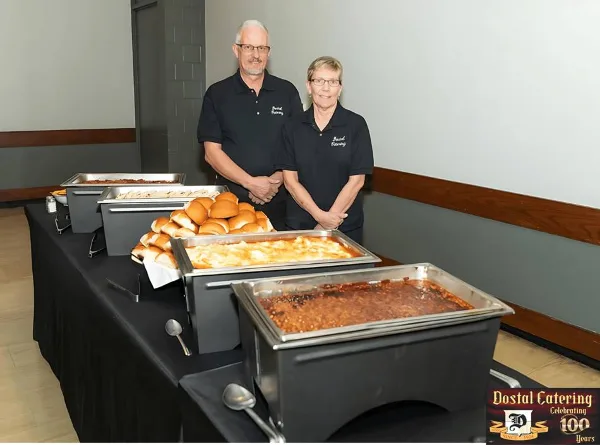Two catering staff members standing behind a buffet table with chafing dishes of food and rolls.