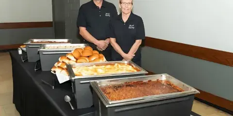 Two catering staff members standing behind a buffet table with chafing dishes of food and rolls.