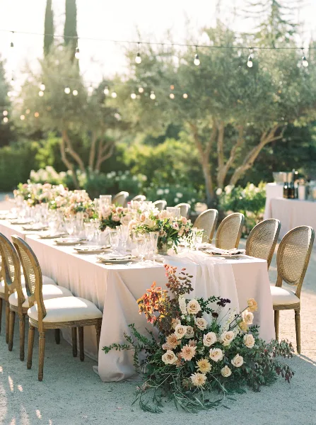 An outdoor wedding reception table with a long white runner, glassware, and lush, organic floral centerpieces and floor arrangements.
