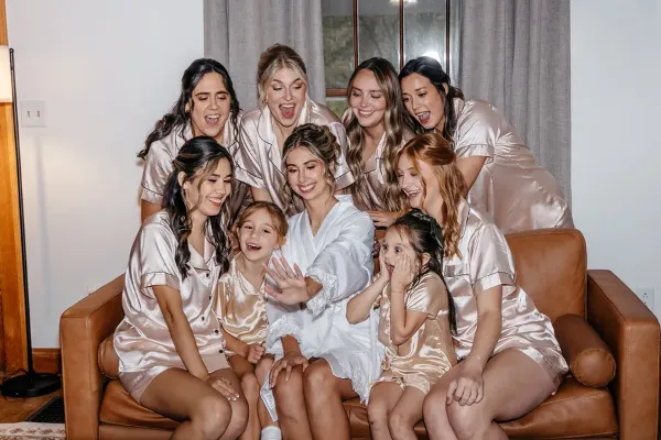 A bride and her bridesmaids in matching satin robes posing together on a leather sofa in a modern room.
