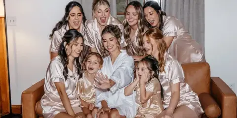 A bride and her bridesmaids in matching satin robes posing together on a leather sofa in a modern room.
