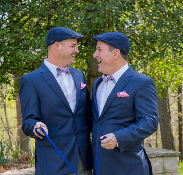 Two smiling men in navy suits, bow ties, and flat caps standing outdoors in a wooded area.