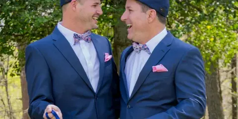 Two smiling men in navy suits, bow ties, and flat caps standing outdoors in a wooded area.