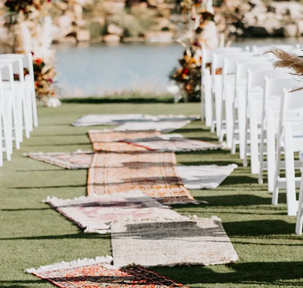 Outdoor wedding ceremony aisle with white chairs and a runner of layered vintage rugs.