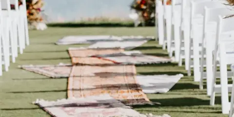 Outdoor wedding ceremony aisle with white chairs and a runner of layered vintage rugs.