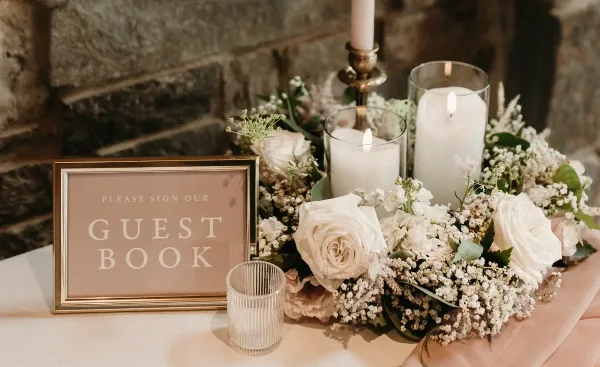 A wedding table setting featuring white roses, baby's breath, lit pillar candles in glass holders, a brass candlestick, and a framed 'Please Sign Our Guest Book' sign against a stone wall.