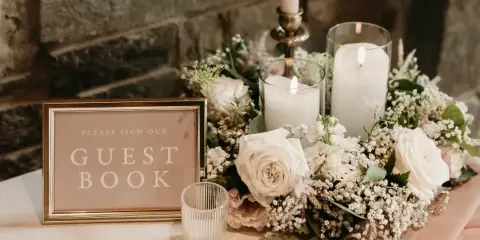 A wedding table setting featuring white roses, baby's breath, lit pillar candles in glass holders, a brass candlestick, and a framed 'Please Sign Our Guest Book' sign against a stone wall.