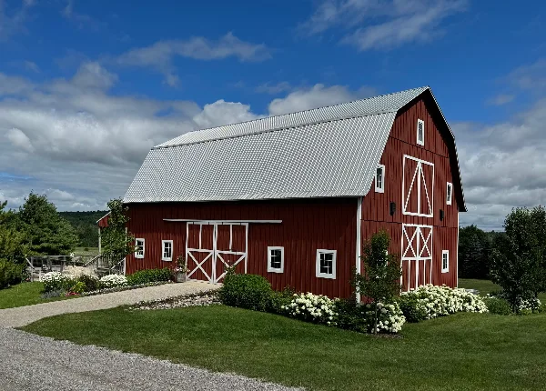 Exterior view of a large red barn with white trim and a metal roof, surrounded by green landscaping and white flowers.