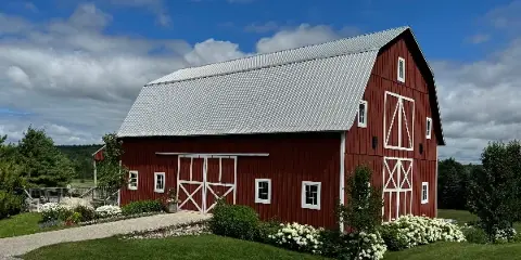 Exterior view of a large red barn with white trim and a metal roof, surrounded by green landscaping and white flowers.