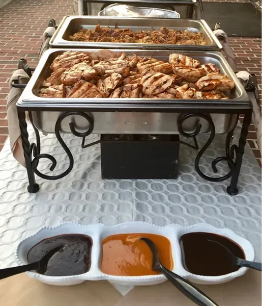 A catering station featuring two chafing dishes filled with grilled meats and a tray of dipping sauces.