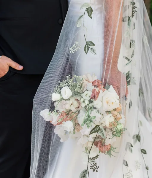 Bride holding a white and blush wedding bouquet with greenery, partially covered by an embroidered veil.