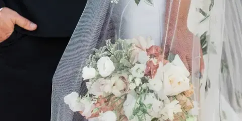 Bride holding a white and blush wedding bouquet with greenery, partially covered by an embroidered veil.