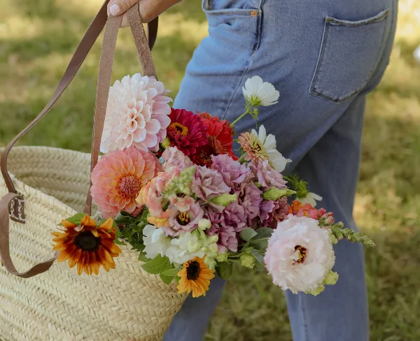 A person in denim jeans holding a woven basket filled with a colorful arrangement of fresh cut flowers including dahlias and sunflowers.