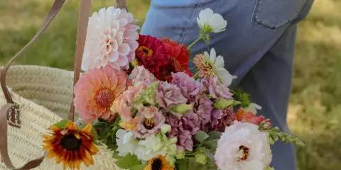 A person in denim jeans holding a woven basket filled with a colorful arrangement of fresh cut flowers including dahlias and sunflowers.