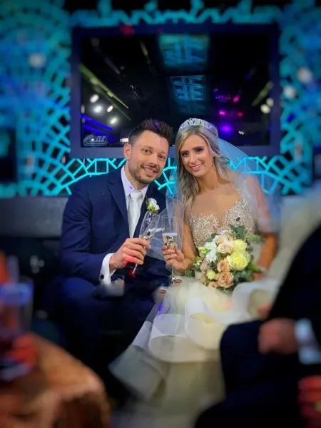 A bride and groom in wedding attire holding champagne flutes and a bouquet.