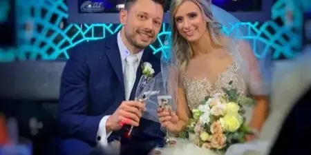 A bride and groom in wedding attire holding champagne flutes and a bouquet.