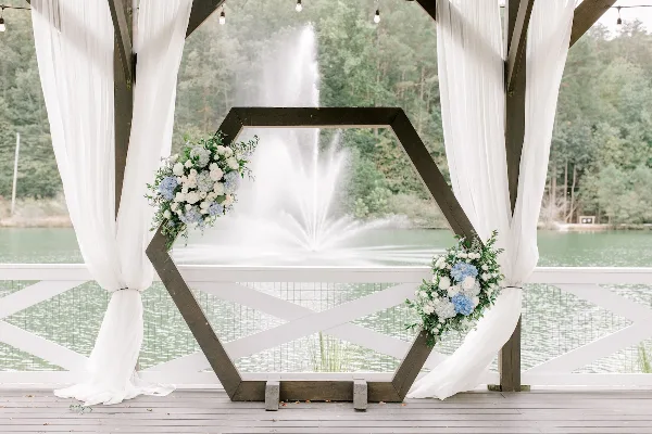 A wooden hexagonal wedding arch decorated with white and blue flowers and white drapery, set on a deck overlooking a lake with a fountain.