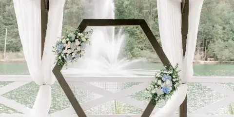 A wooden hexagonal wedding arch decorated with white and blue flowers and white drapery, set on a deck overlooking a lake with a fountain.