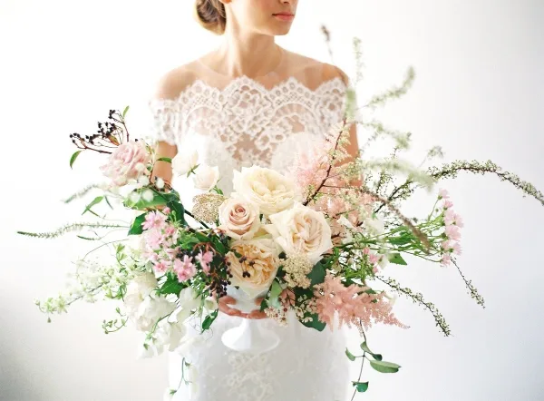 A bride in a white lace gown holding a lush, romantic bridal bouquet featuring cream roses, pink astilbe, and greenery.