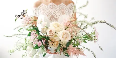 A bride in a white lace gown holding a lush, romantic bridal bouquet featuring cream roses, pink astilbe, and greenery.