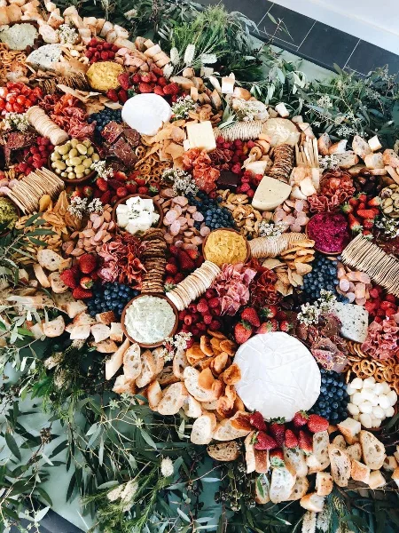 Overhead view of a large catering spread featuring cheeses, cured meats, fresh berries, bread, and greenery arranged on a table.