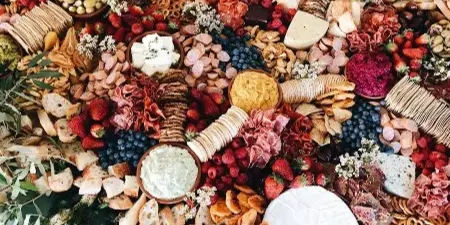 Overhead view of a large catering spread featuring cheeses, cured meats, fresh berries, bread, and greenery arranged on a table.