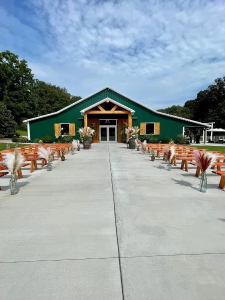 Exterior view of a green barn wedding venue with a concrete patio and rows of wooden benches set up for a ceremony.