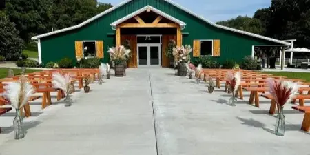 Exterior view of a green barn wedding venue with a concrete patio and rows of wooden benches set up for a ceremony.