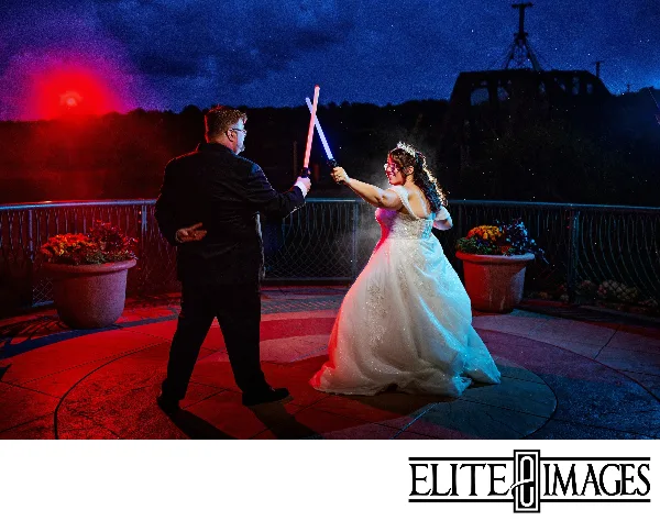 A bride and groom holding crossed light sabers on a rooftop terrace at night.