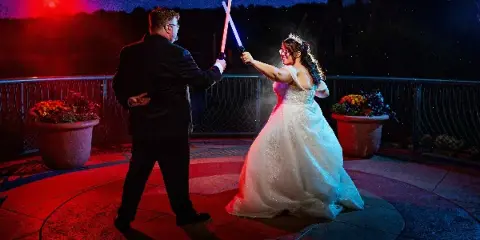 A bride and groom holding crossed light sabers on a rooftop terrace at night.