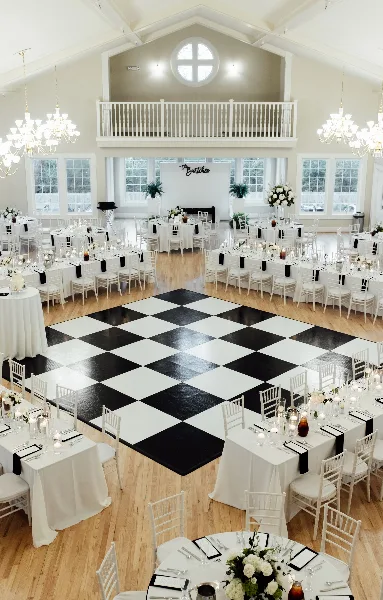 A wedding reception hall set up with white tables, chairs, and a large black-and-white checkered dance floor.
